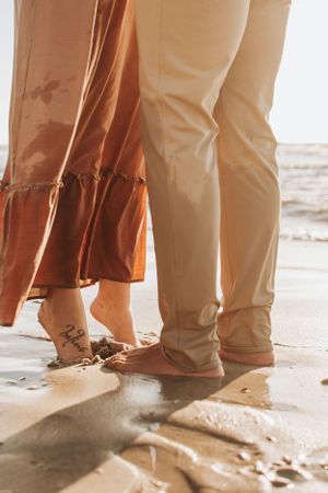 Feet in casual footwear standing on a sandy beach during golden hour.