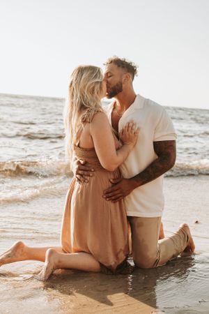 Couple sitting together at the water's edge on a sandy beach at sunset.