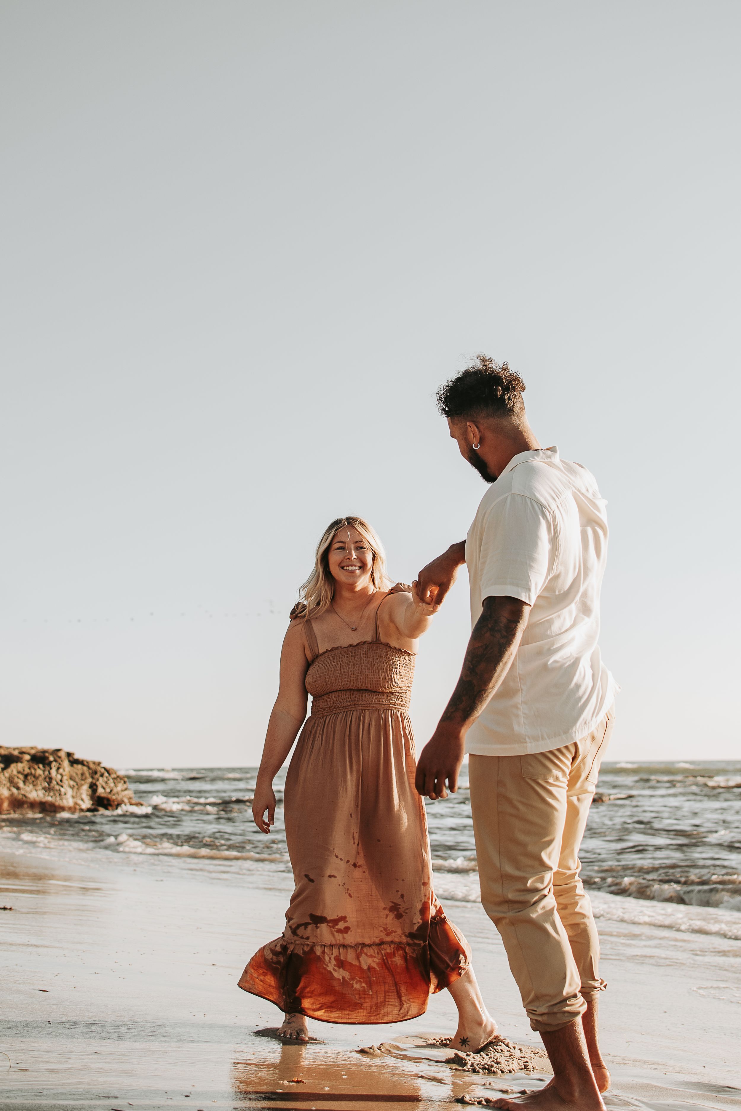 A couple walks along the beach together at sunset near rocky coastal formations.