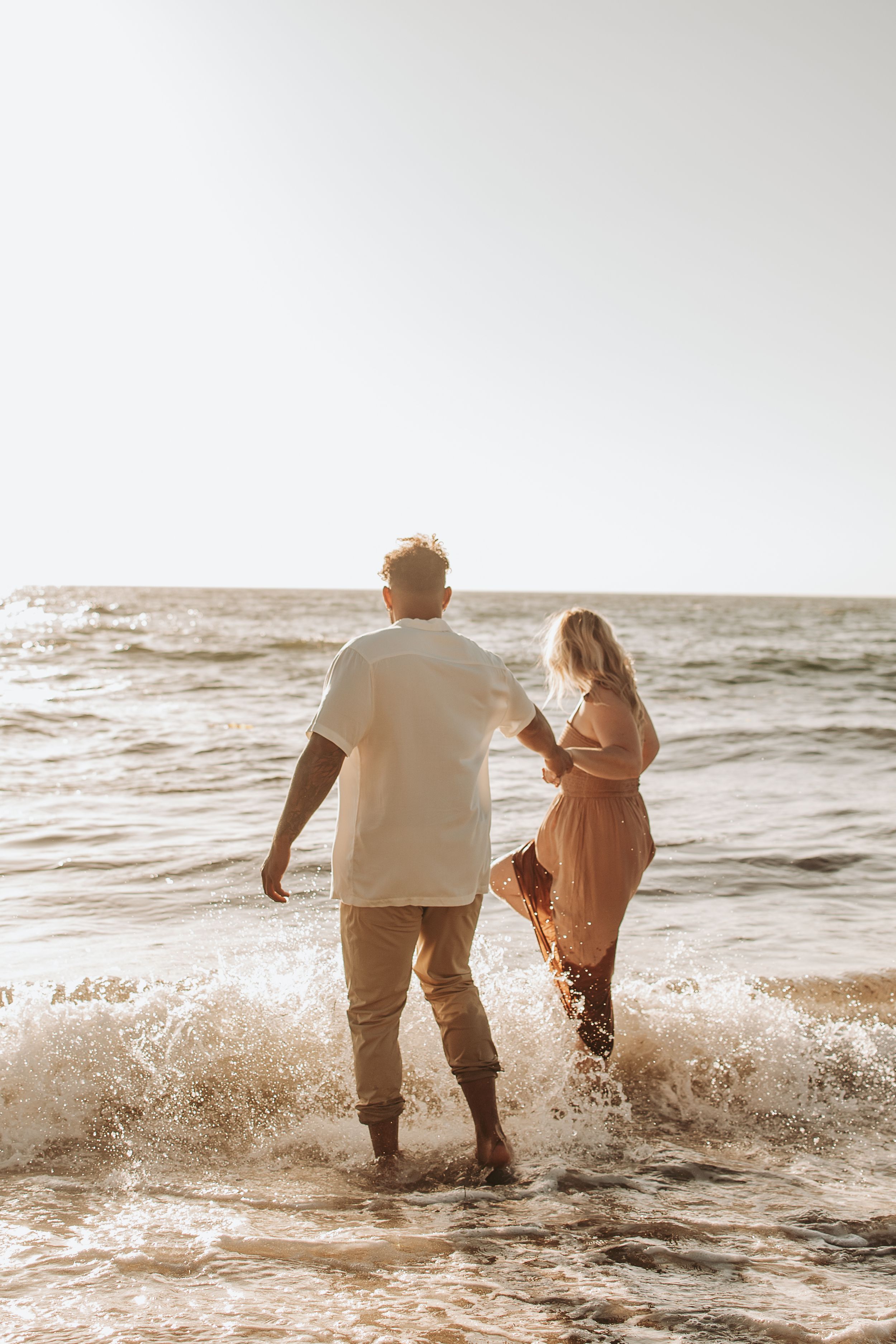 A couple wades through crashing ocean waves together during golden hour.