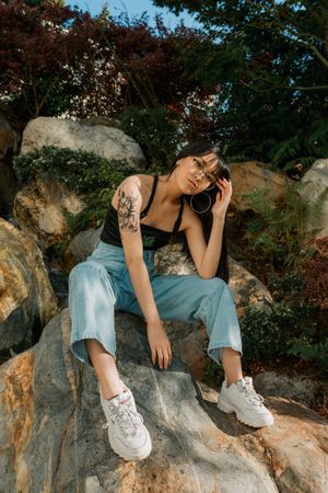 Artistic portrait on rocky terrain with chunky white sneakers, light jeans, and dark top against lush foliage background.