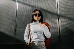 A person wearing a light gray sweater, sunglasses and crossbody bag poses against a metallic backdrop.