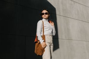 Stylish person in sunglasses wearing neutral outfit with brown suede bag poses against gray wall in natural lighting.