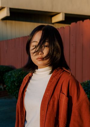 Person wearing a white turtleneck and rust-colored button-up jacket poses against a colorful fence in warm sunset lighting.