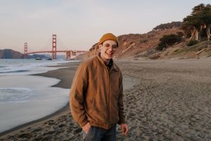 Someone in a brown jacket and hat stands on Baker Beach with the Golden Gate Bridge visible in the background at sunset.