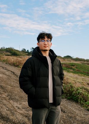 Someone wearing a black puffer jacket and glasses stands on a dirt path against a cloudy sky and grassy hillside.