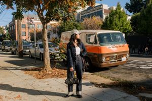 A person in black attire stands on a sidewalk with an orange vintage van parked in the background on a sunny autumn day.