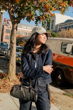 Street style fashion shot with denim outfit, baseball cap and textured handbag amid autumn trees and urban setting.