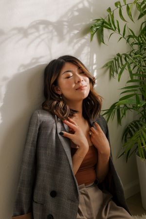 Thoughtful woman in gray blazer and brown top poses against white wall with tropical plant shadows.