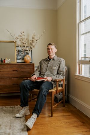 A relaxed scene showing someone in a chair reading near a window, with a wooden dresser and floral decor in the corner.