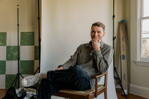 A casual portrait of someone sitting in a vintage wooden chair against a wall with green checkered tiles.