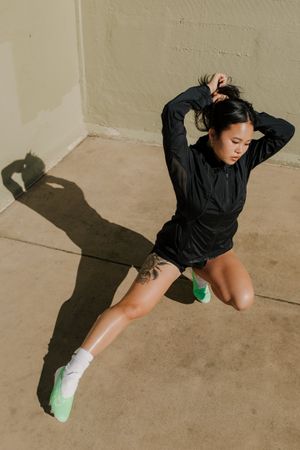 Athletic woman in black jacket and shorts jumping against beige wall casting dramatic shadow.