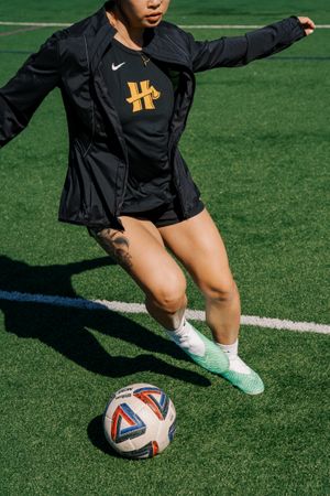 Soccer player in black Nike jacket and shorts dribbles ball on artificial turf field with bright sunlight casting shadow.