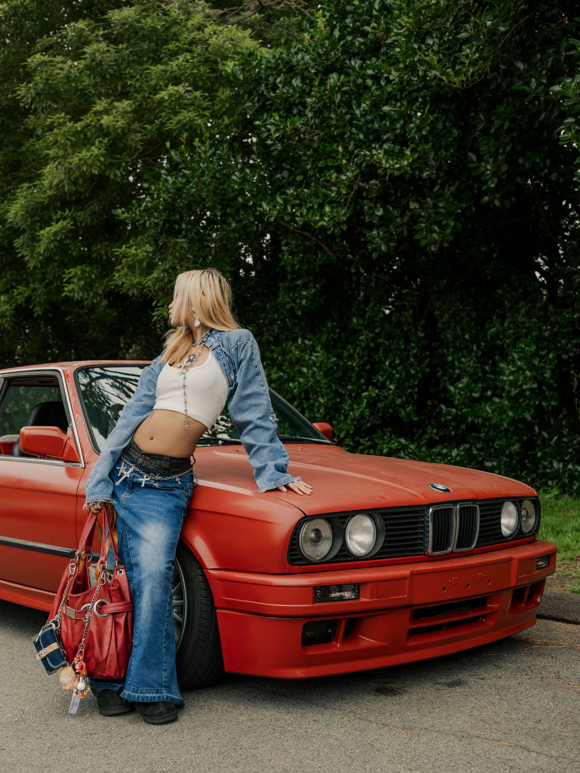 A vintage red BMW E30 M3 with someone in casual denim attire leaning against it near green trees.