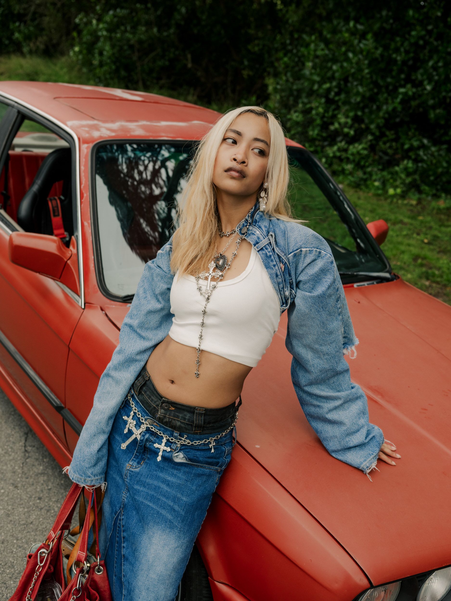 Young model leaning against red vintage car wearing denim jacket, cropped top and jeans in outdoor photoshoot.