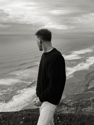 A person in black sweater and white pants stands on coastal cliffs overlooking the ocean in a moody black and white scene.