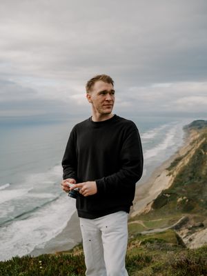 Someone in a black sweater and white pants stands on a scenic coastal cliff overlooking the ocean under cloudy skies.