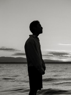 A silhouette of a figure stands contemplatively by the ocean at sunset in black and white.