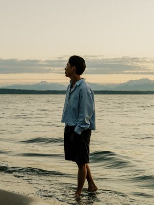 A person in a light blue shirt stands ankle-deep in the water at sunset, with mountains visible on the horizon.