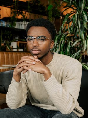 A person in a cream sweater smiles thoughtfully while seated in a cozy restaurant setting with greenery in the background.