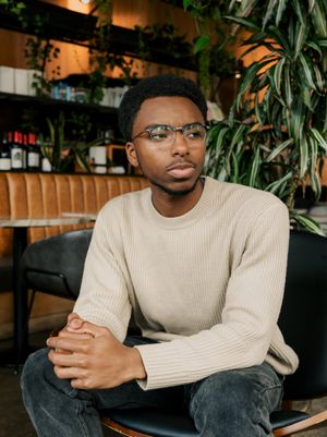 Person in a cream sweater sits thoughtfully in an indoor space with tropical plants and warm lighting in the background.