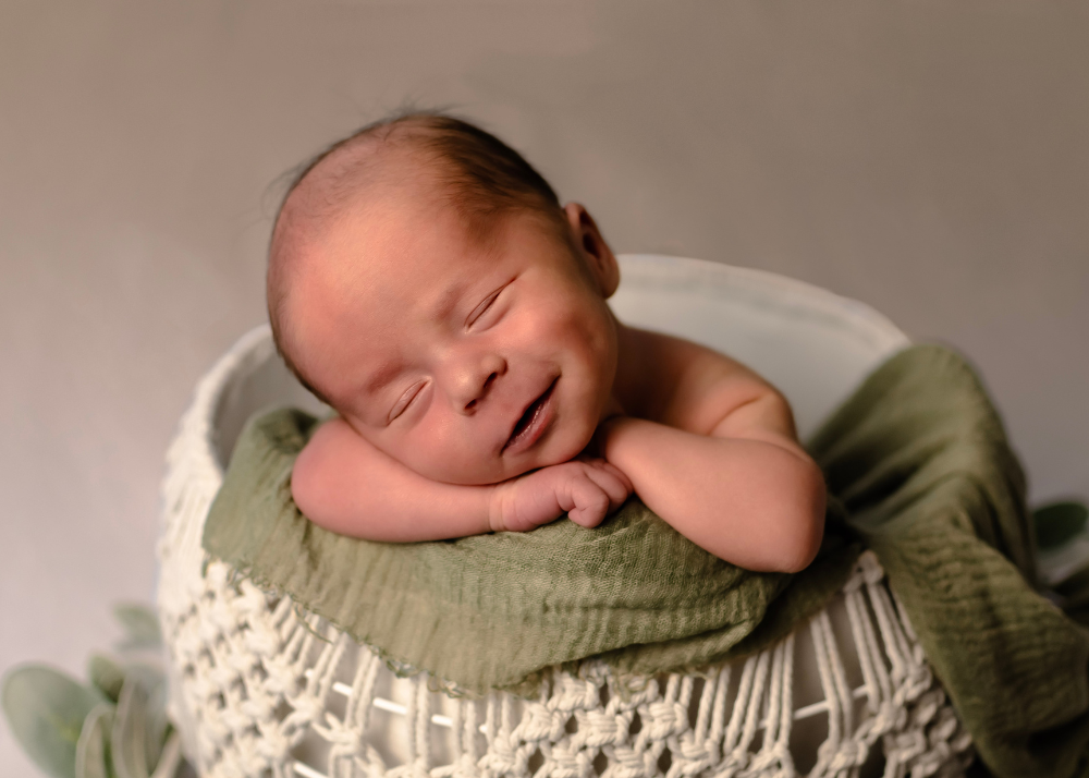 A sleeping baby wearing olive green clothing rests peacefully in a textured cream-colored basket.