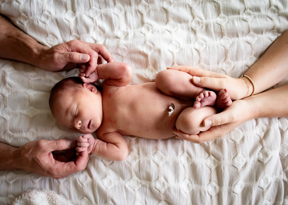 Adult hands tenderly hold a sleeping newborn baby lying on white textured bedding.