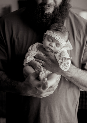 A black and white photograph showing a newborn baby wearing a knit hat being cradled in strong tattooed arms.