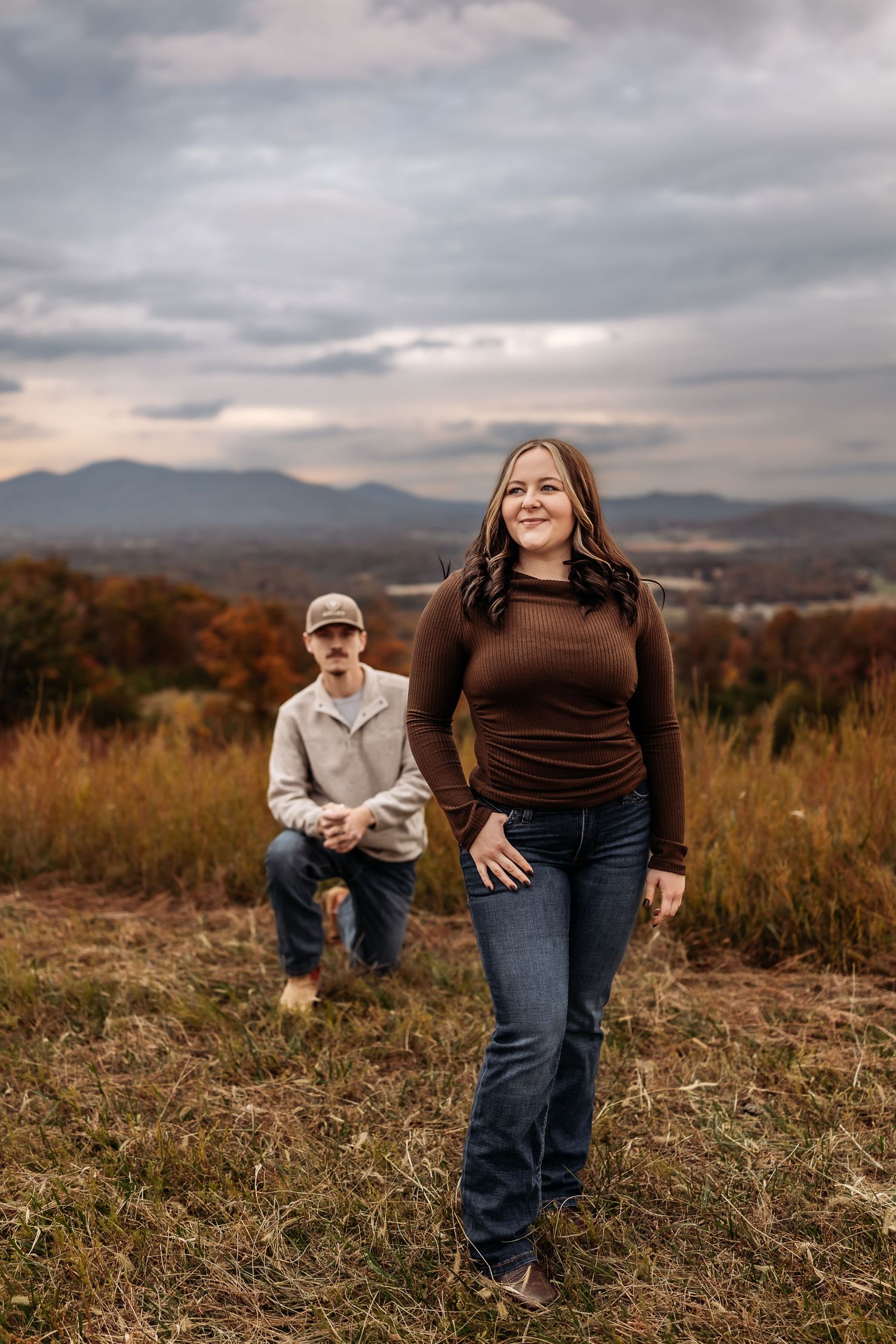 Two people pose on a hilltop during autumn with mountains and cloudy skies in the background.