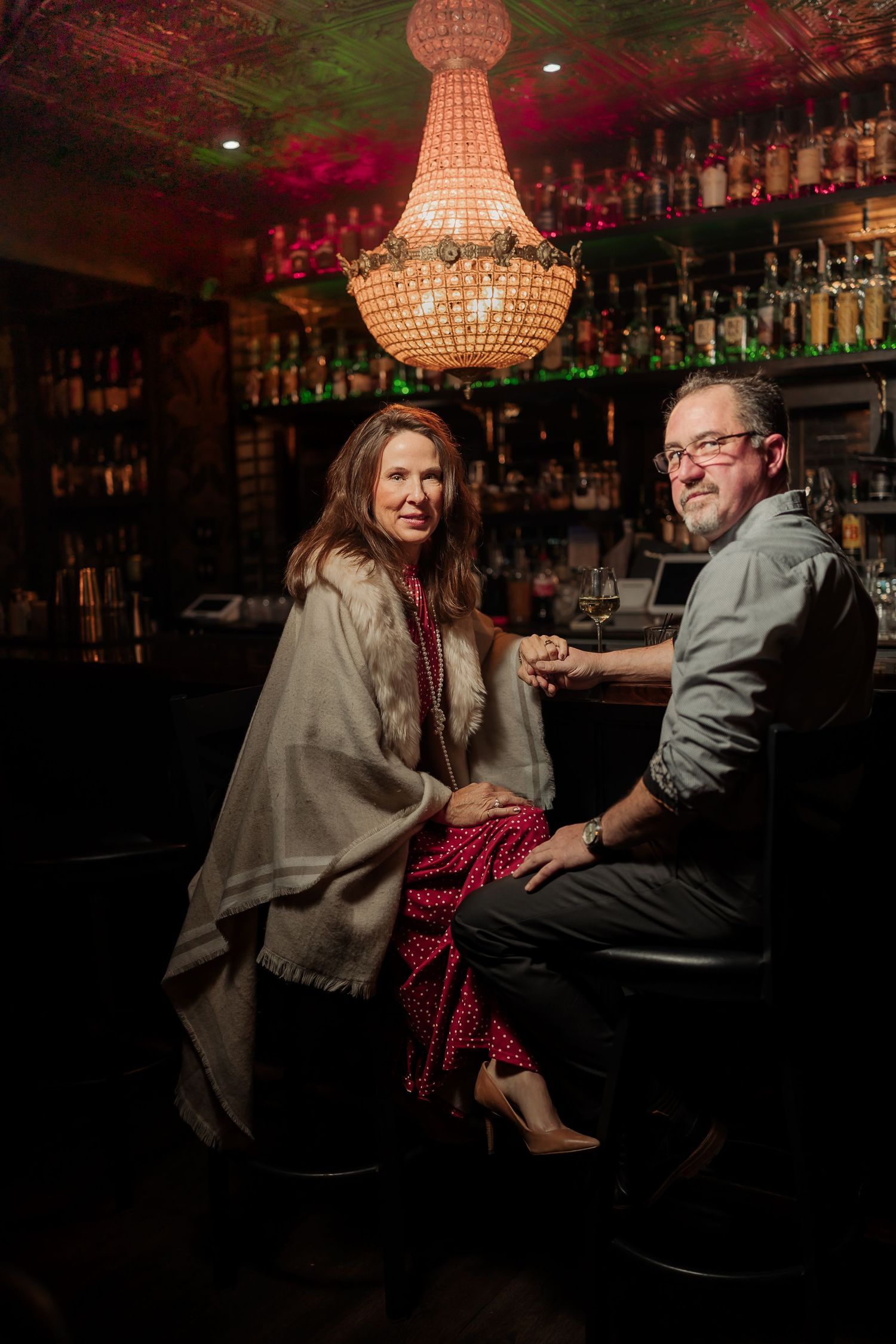 Two people sit intimately at a dimly lit bar with crystal chandeliers overhead creating a romantic ambiance.