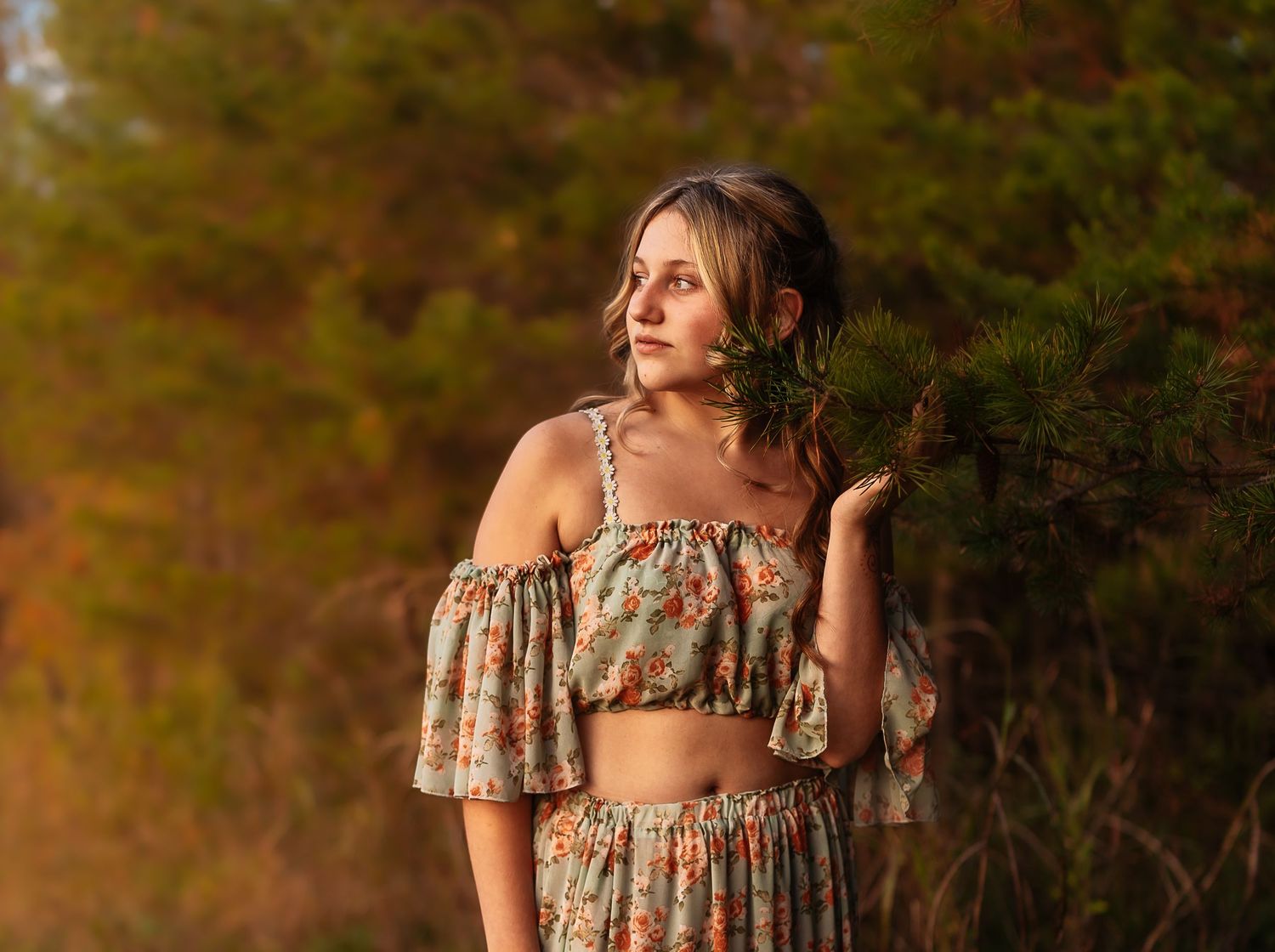 A person in a floral dress poses outdoors during golden hour with sunlit trees and foliage in the background.