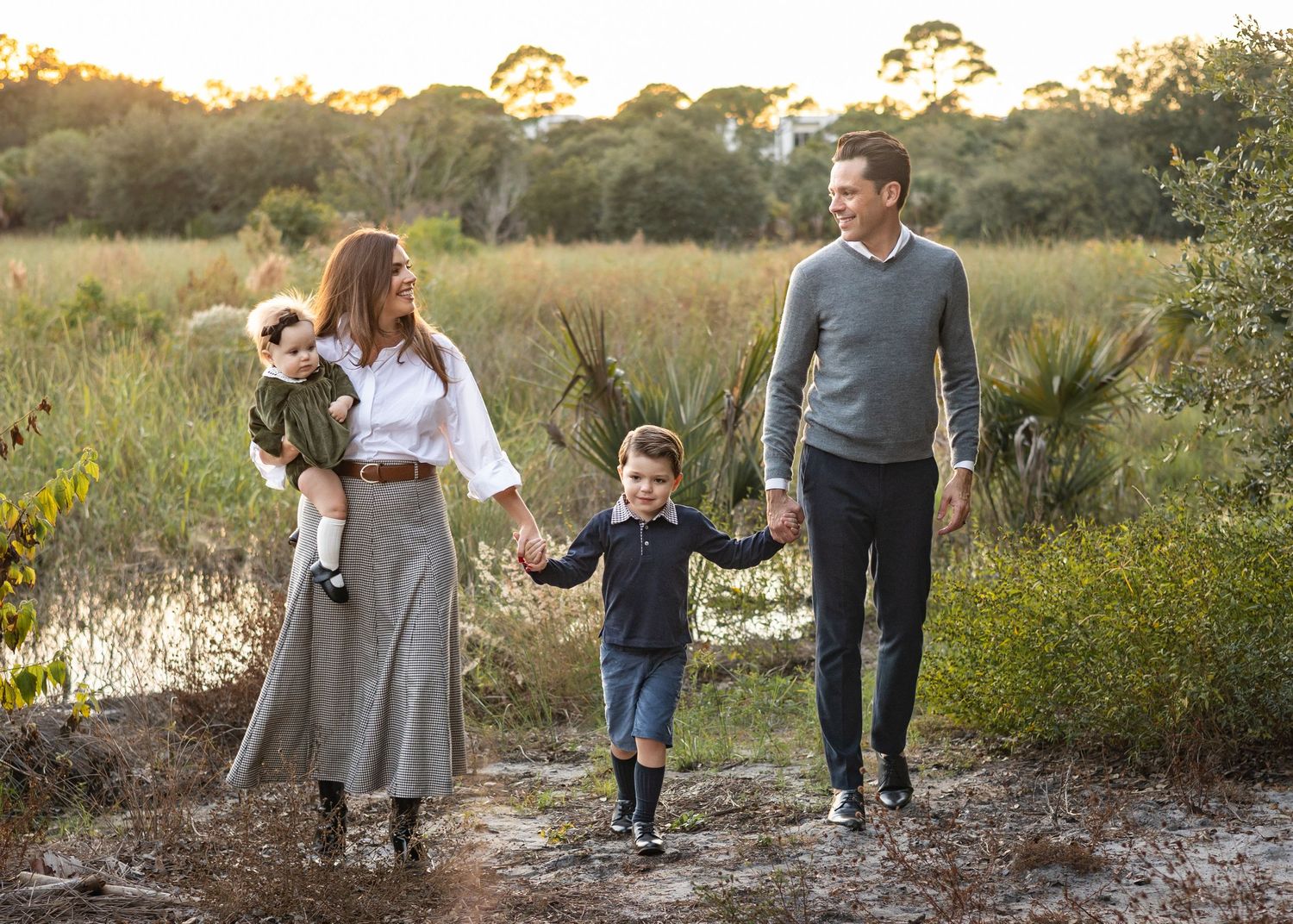 A couple in olive green and rust colored boho-style attires pose candidly with their daughter on a rocky beach during a sunset photoshoot.