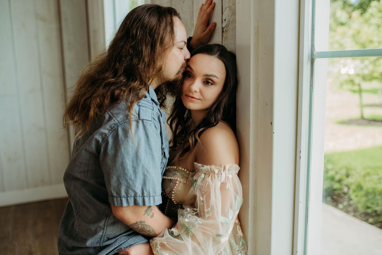 Romantic couple in casual attire embracing near window with natural lighting.