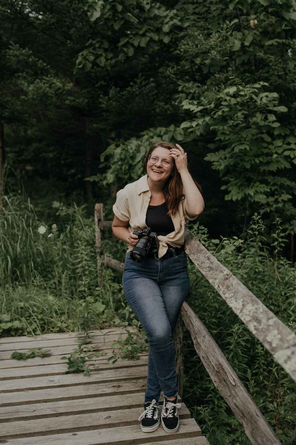 A photographer laughs while posing with her camera on a wooden boardwalk surrounded by greenery.
