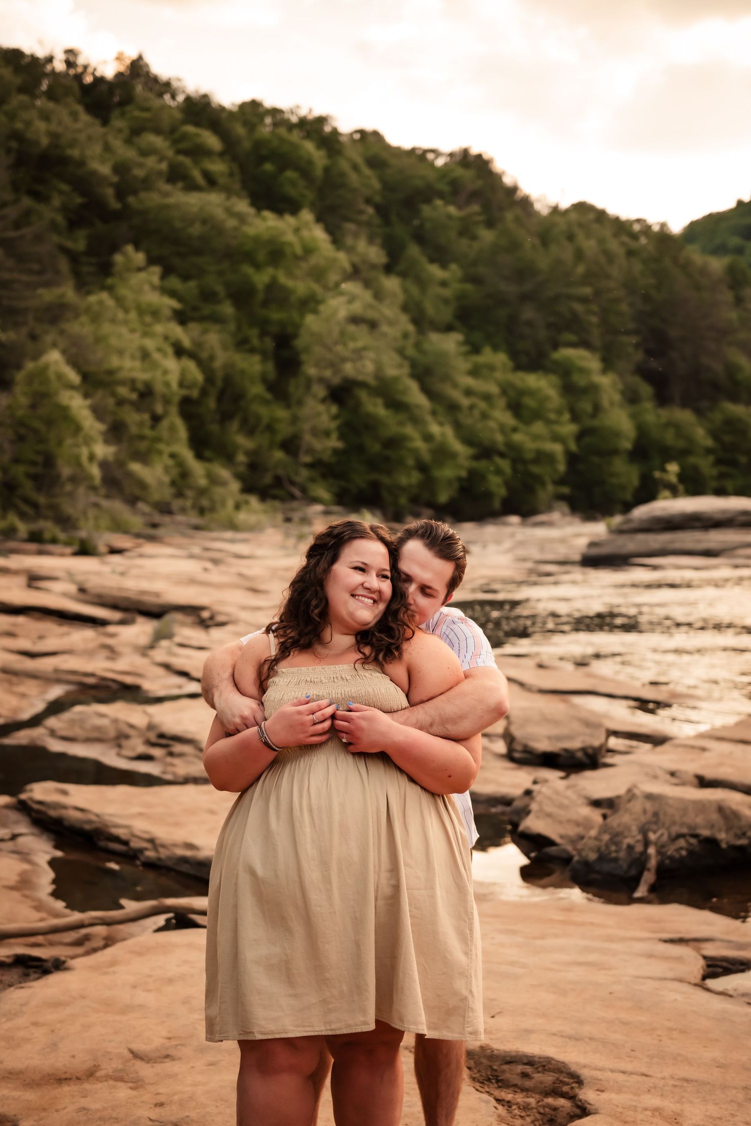 Romantic couple embracing on rocky riverbed with forested hillside in background.