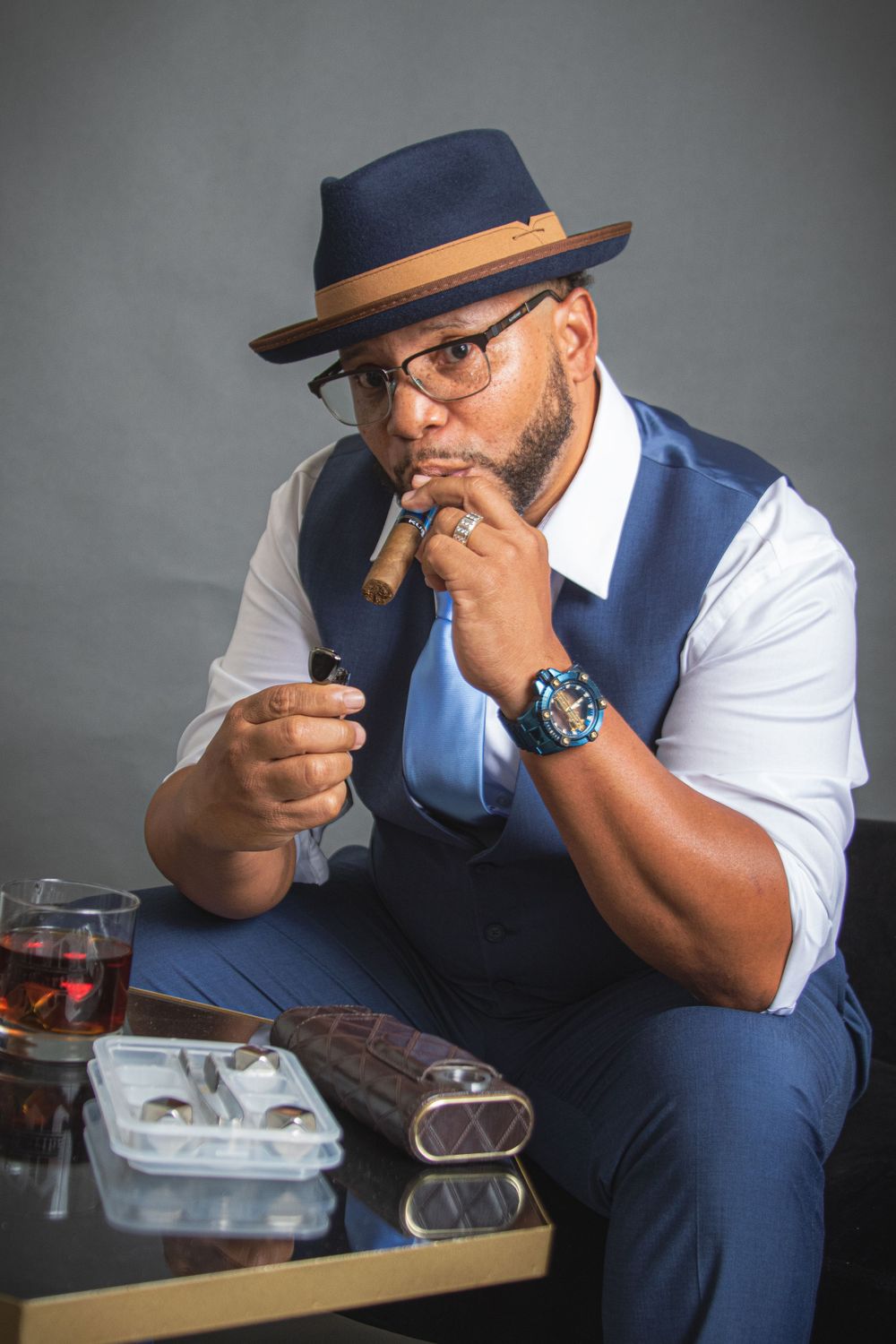 Person in vest and fedora enjoying cigar with drink and accessories on table.