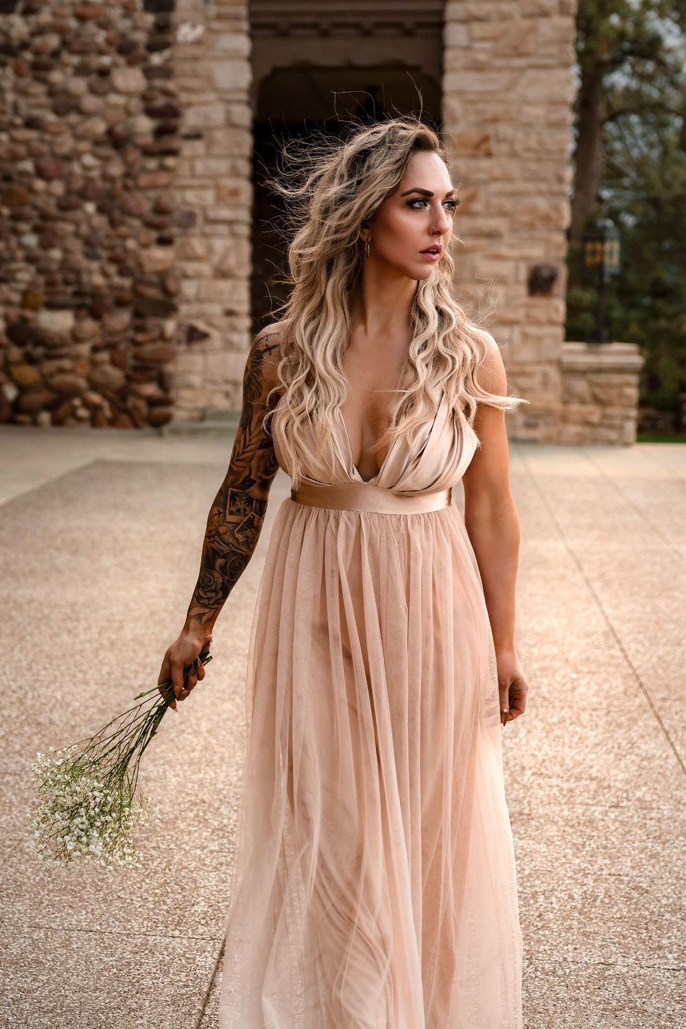 Person in flowing blush gown holds baby's breath flowers near stone architecture.