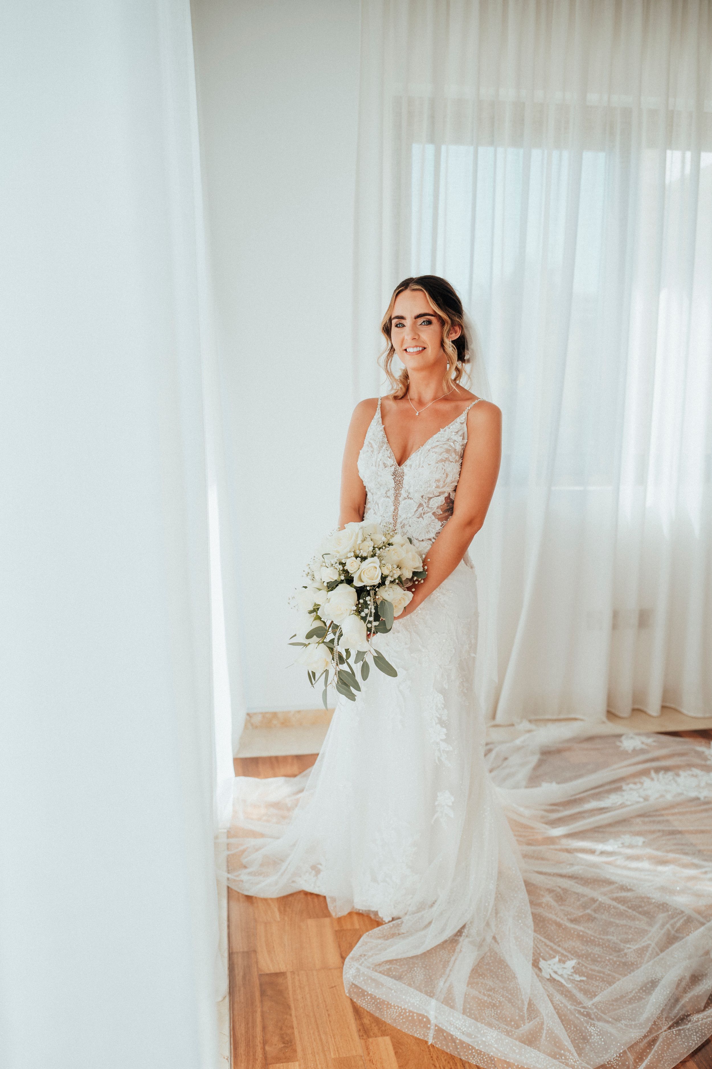 Beautiful bride in lace wedding dress holds white floral bouquet against sheer curtain backdrop in sunlit room.