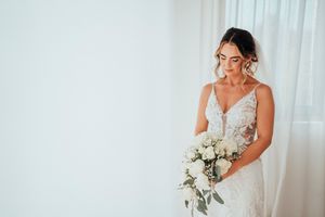A bride in a white wedding dress holds a stunning bouquet of white flowers against a light backdrop.