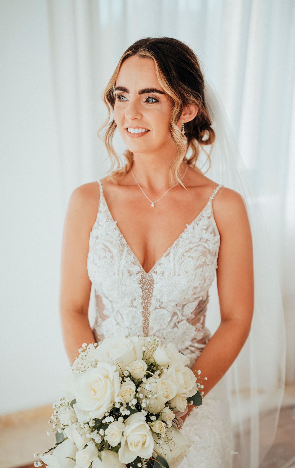A bride in a lace wedding dress holds an elegant white rose bouquet against a light backdrop.