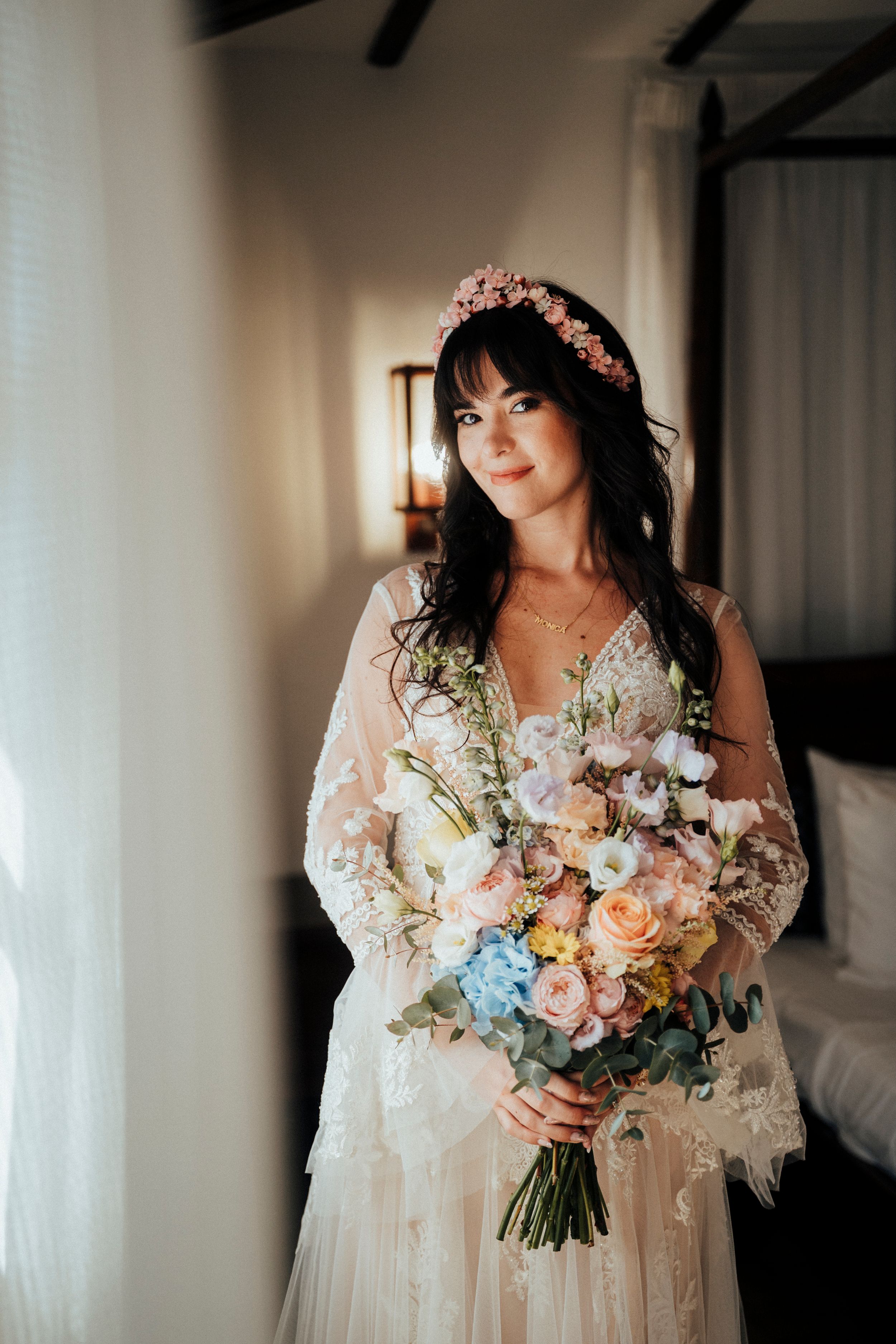 A bride in a lace wedding dress holds a romantic bouquet of pastel roses and delicate flowers in soft lighting.