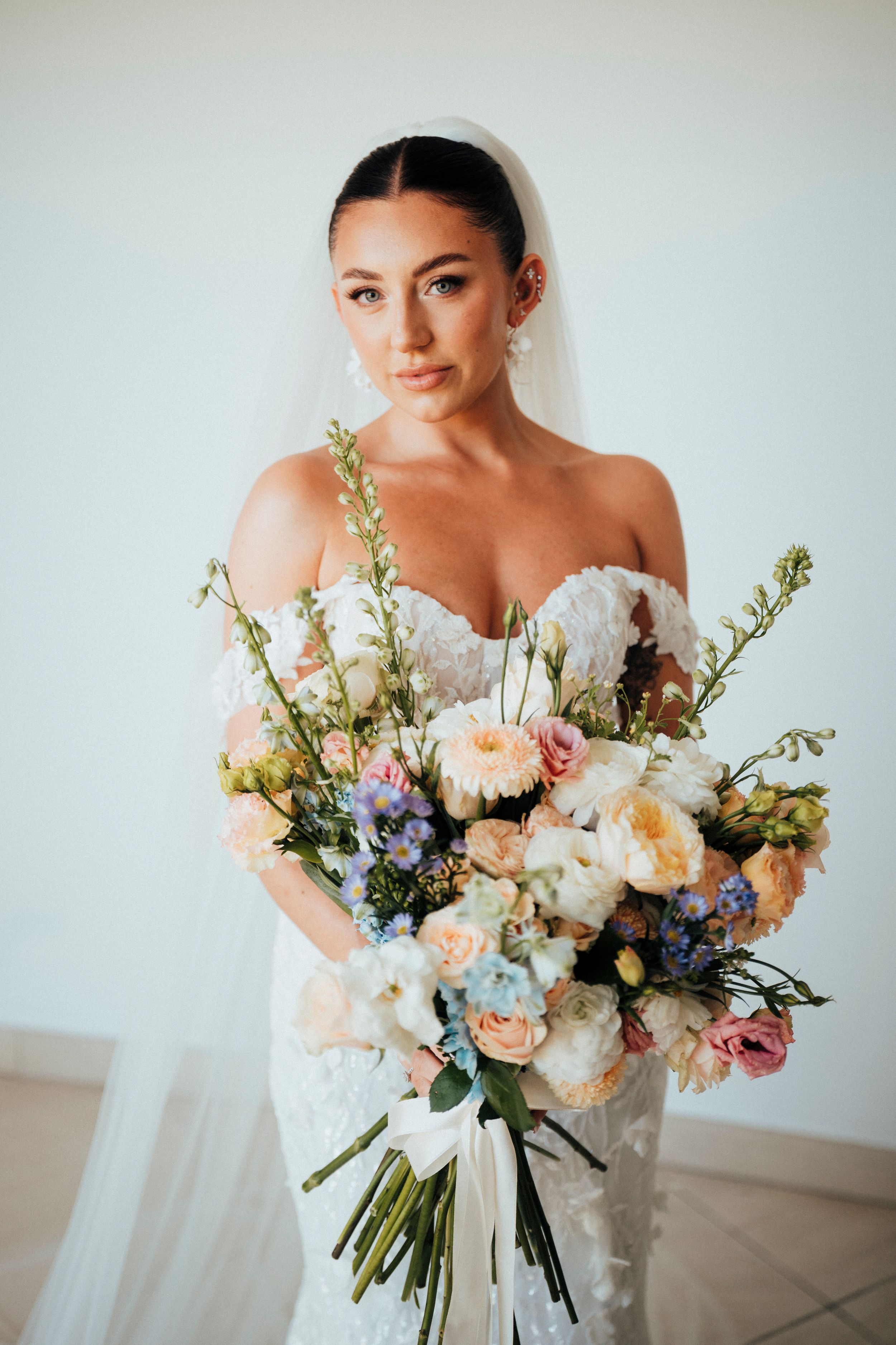 Bride in white strapless gown holds romantic pastel bouquet with peach roses, white peonies and delicate blue flowers.