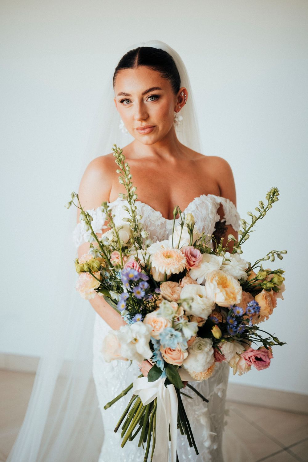 Bride in white strapless gown holds romantic pastel bouquet with peach roses, white peonies and delicate blue flowers.