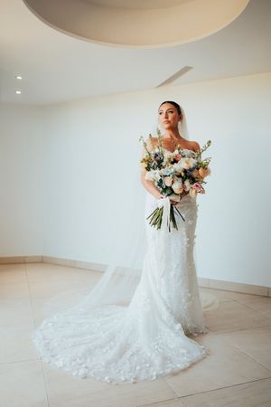 Elegant bride in lace off-shoulder wedding gown with flowing train poses in bright white room with beautiful bouquet.