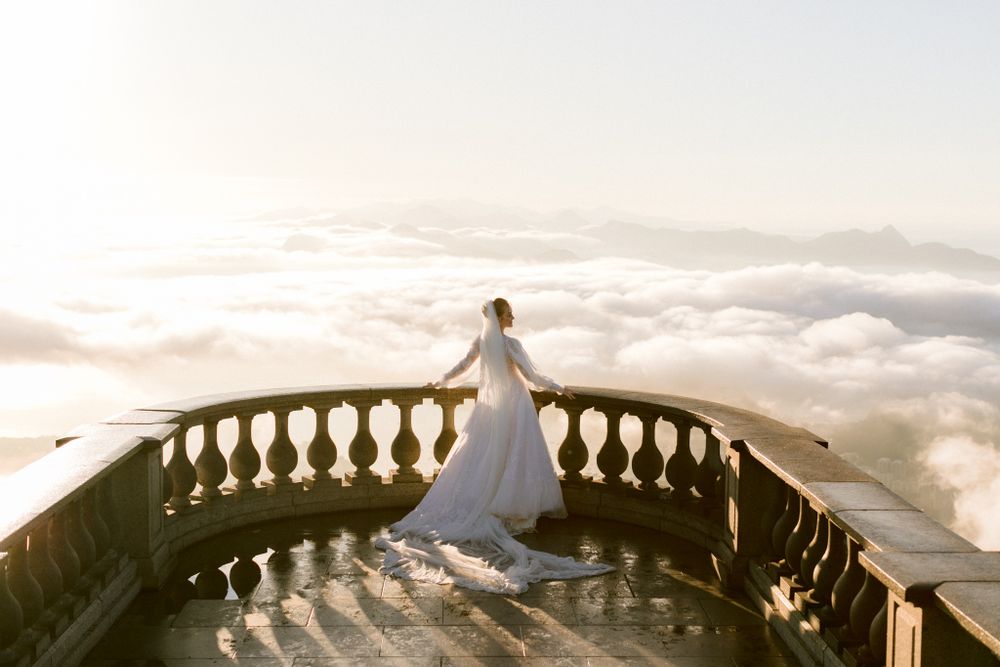 Silhouetted figure in a wedding dress stands on a balcony overlooking a sea of clouds at sunset.