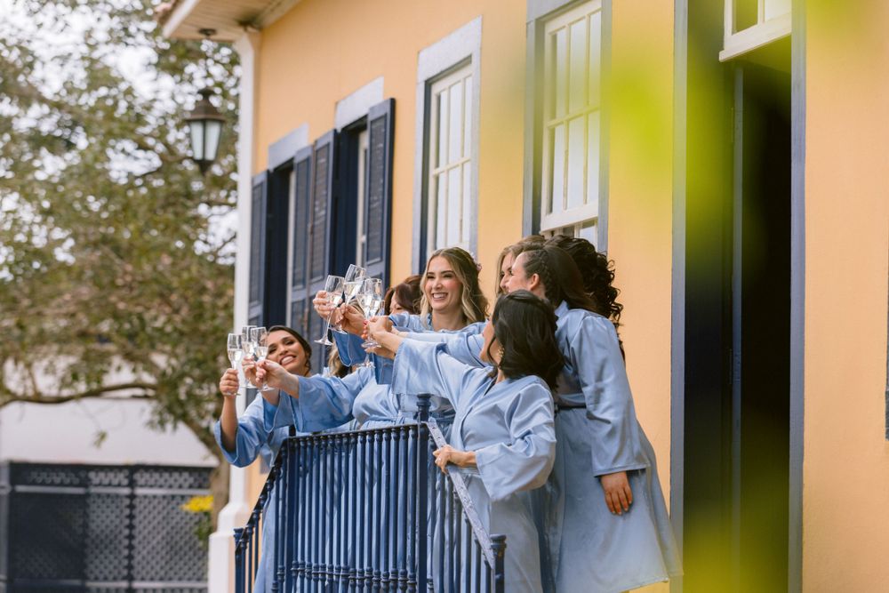 Wedding party in matching blue robes cheers from balcony of yellow building.