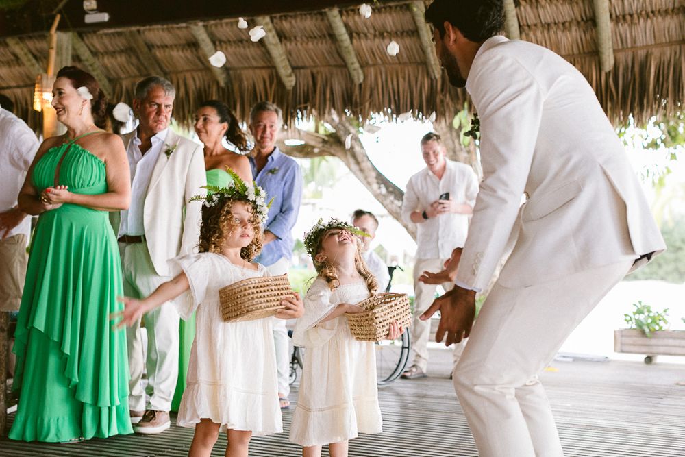 Wedding guests in bright attire dancing under a thatched roof pavilion.