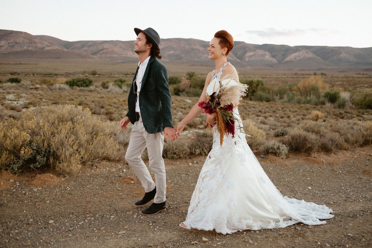 A stylish couple in wedding attire walking through a desert landscape with mountains in the distance.
