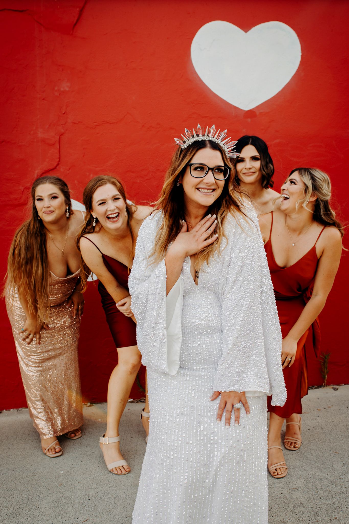 Wedding party poses against red wall with white heart, bride in sequined dress and crown.