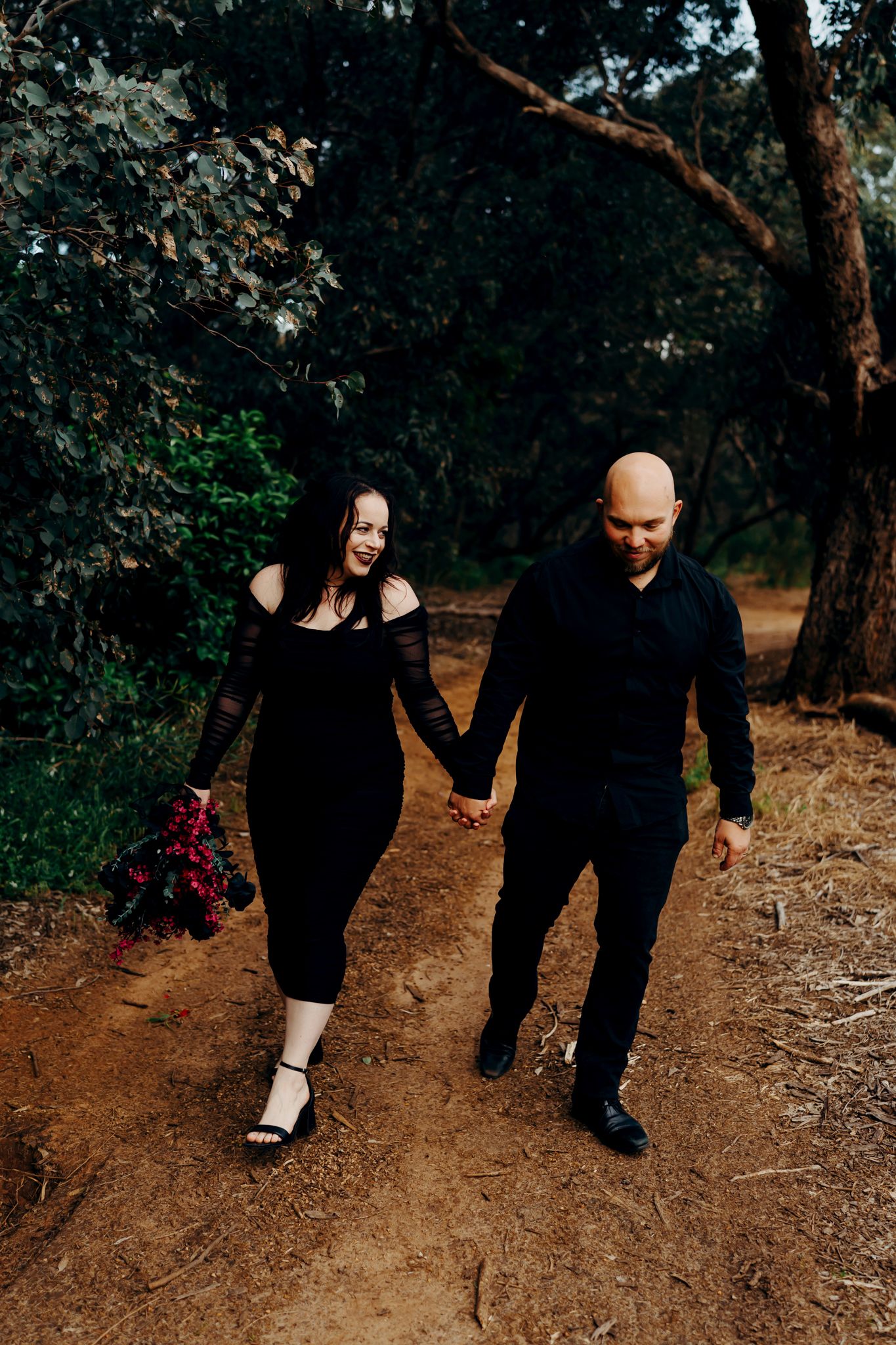 A couple walks together on a dirt path through a wooded area wearing black clothing on an autumn day.